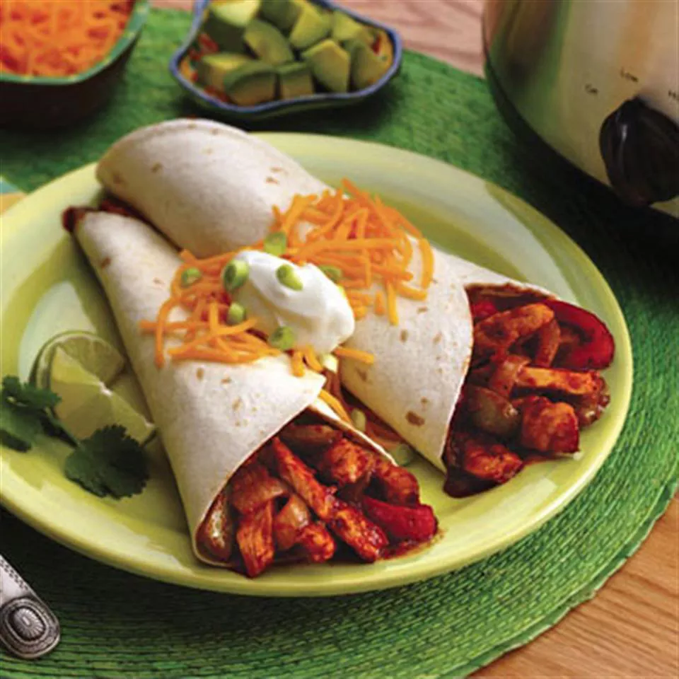 Close up view of slow cooker chicken fajitas in tortillas garnished with cheese, sour cream, green onions, lime wedges and cilantro on a green plate