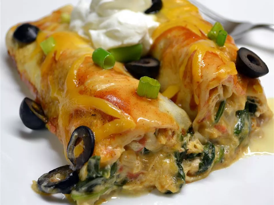Close up view of spinach and chicken enchiladas garnished with sour cream, green onions and black olives on a white plate