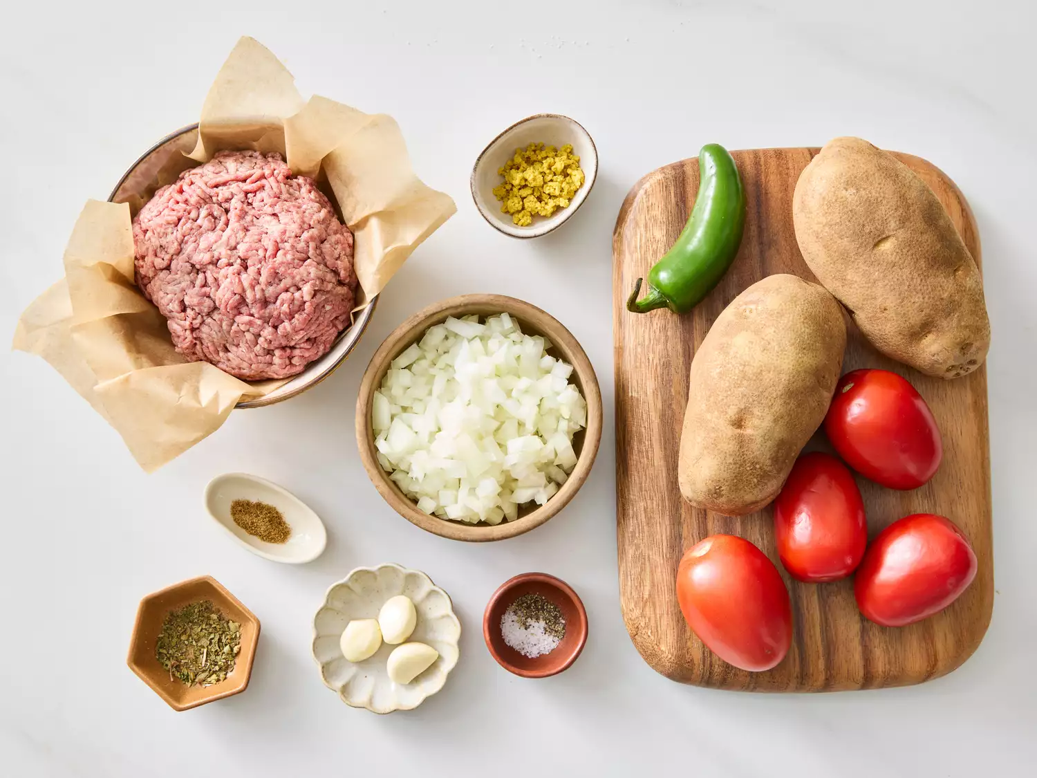 Ground beef, chopped onion, garlic cloves, potatoes, Roma tomatoes, jalapeño, and spices arranged on a surface, preparation for cooking.