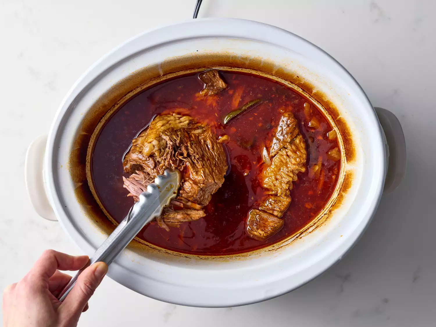 Cooked brisket in slow cooker being lifted with tongs