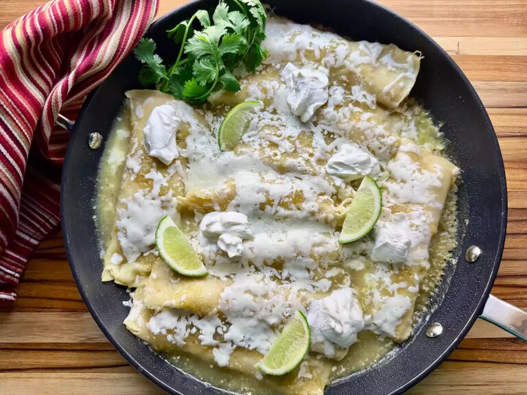 Overhead view of green chicken enchiladas with cheese and lime wedges in skillet