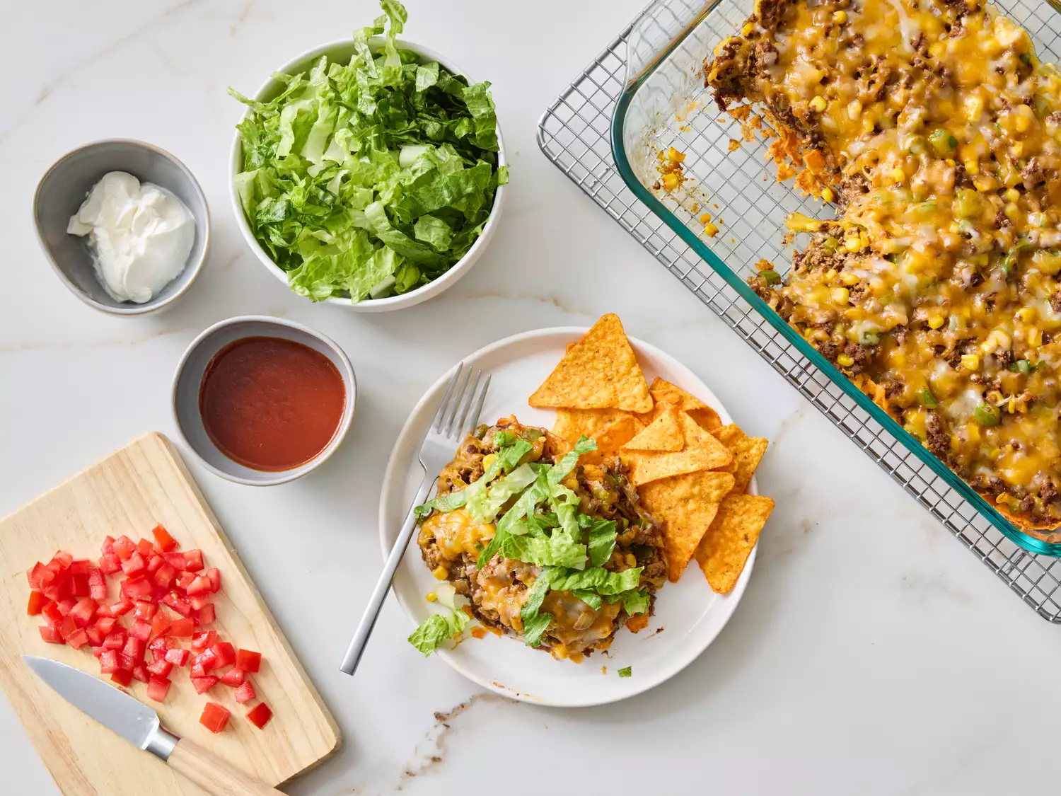 Dish featuring taco salad casserole served with tortilla chips, garnished with lettuce and tomatoes, additional toppings on the side