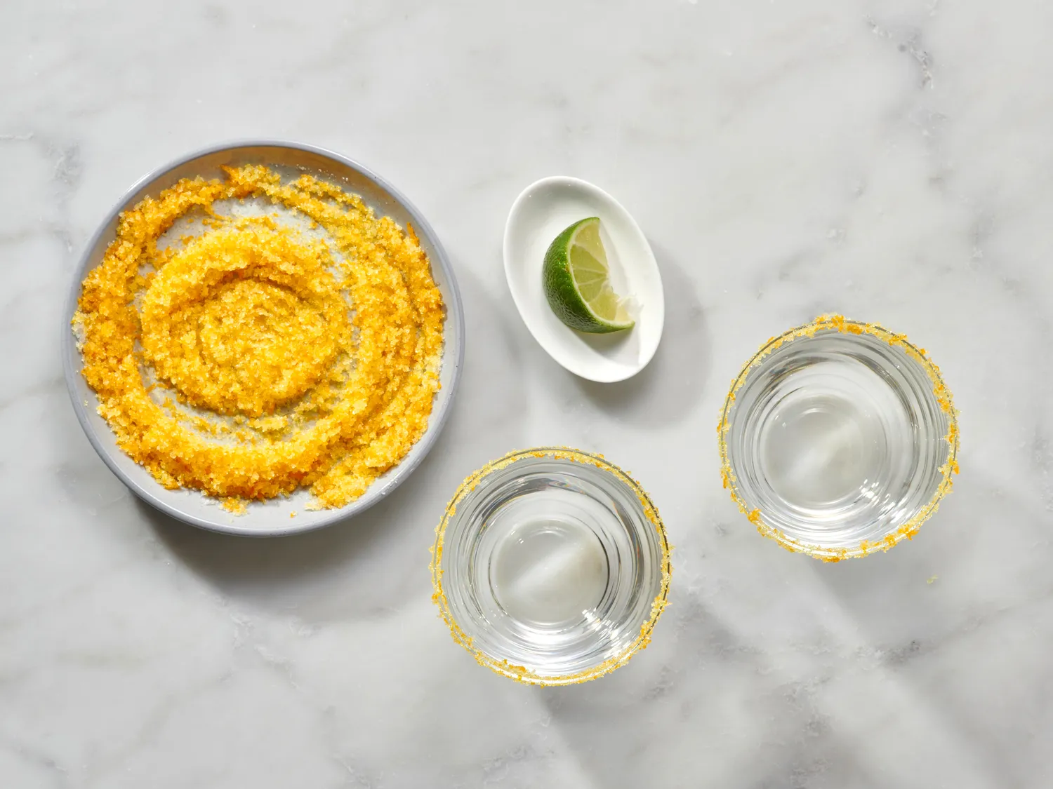 Rimmed glasses next to lime slices and a plate with orange sugar mixture