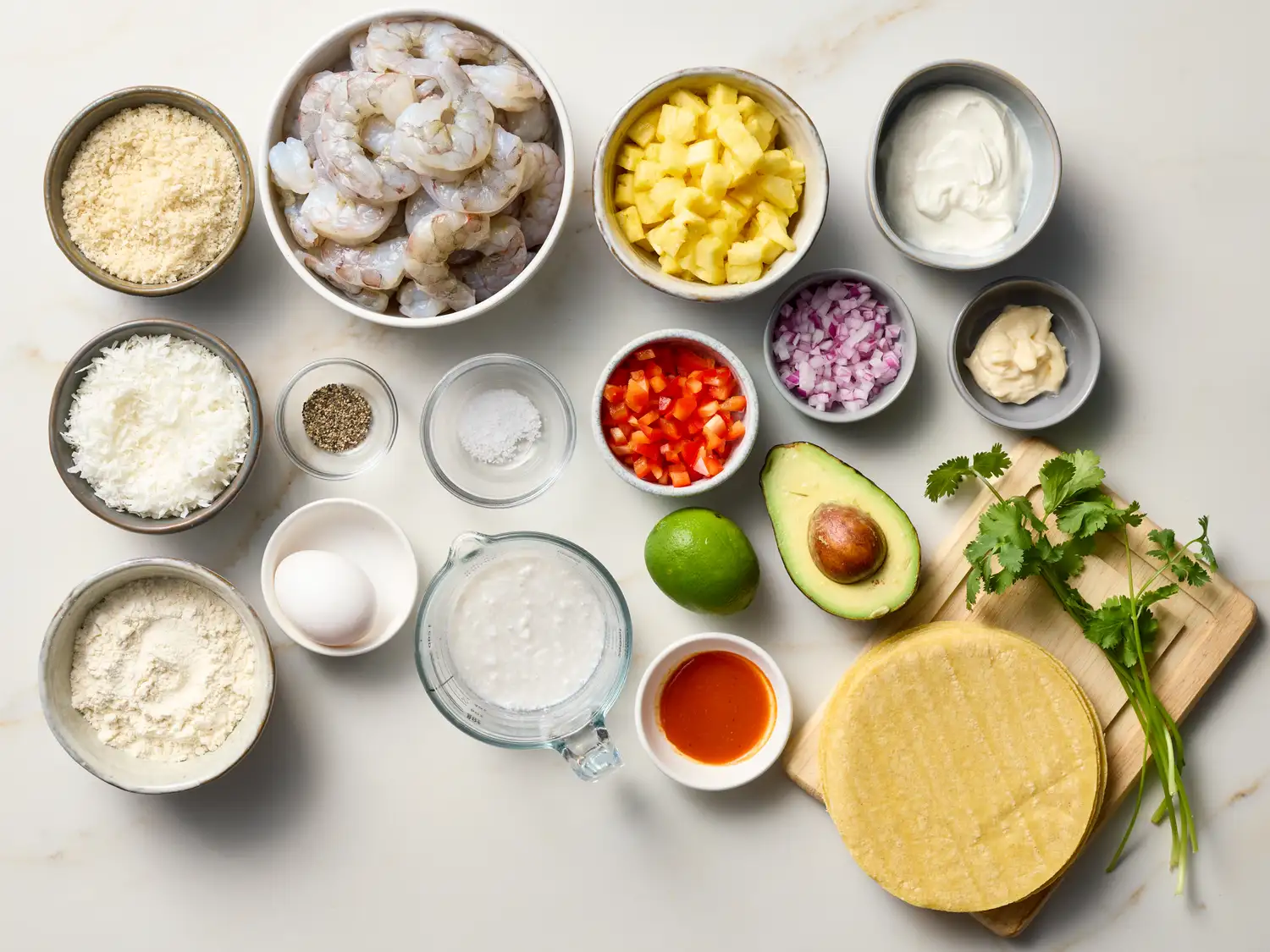Ingredients for crispy coconut shrimp tacos arranged on a surface, including shrimp, coconut, spices, avocado, tortillas, and toppings