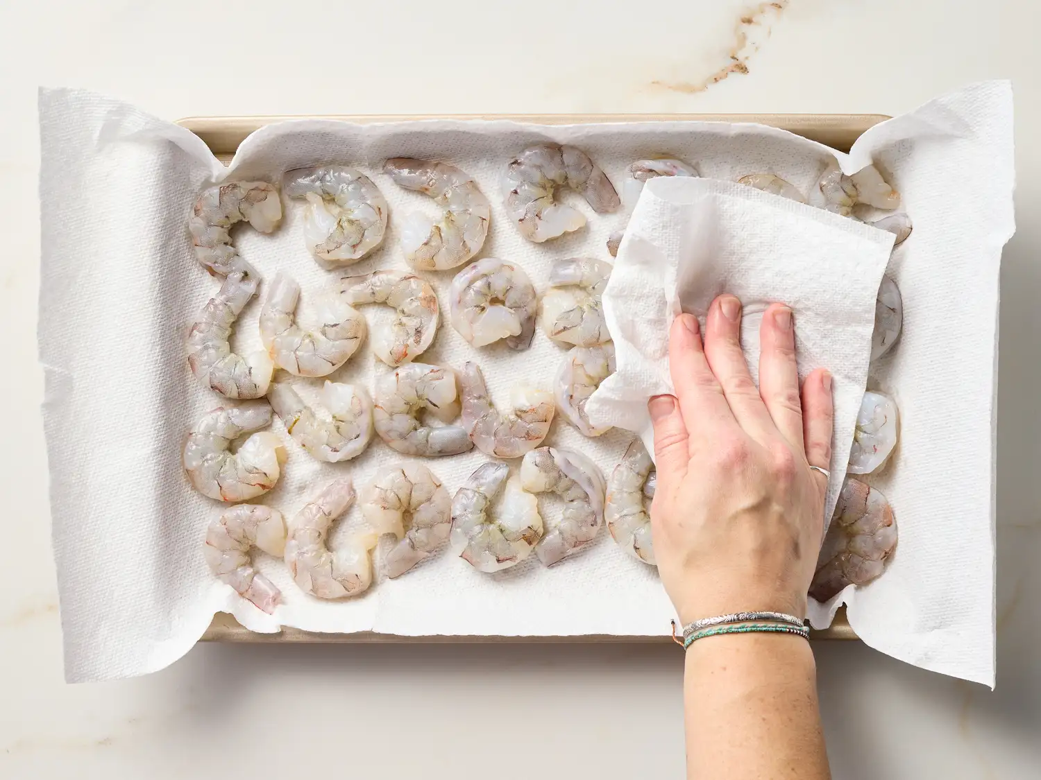 Person blotting raw shrimp with a paper towel on a tray lined with paper towels
