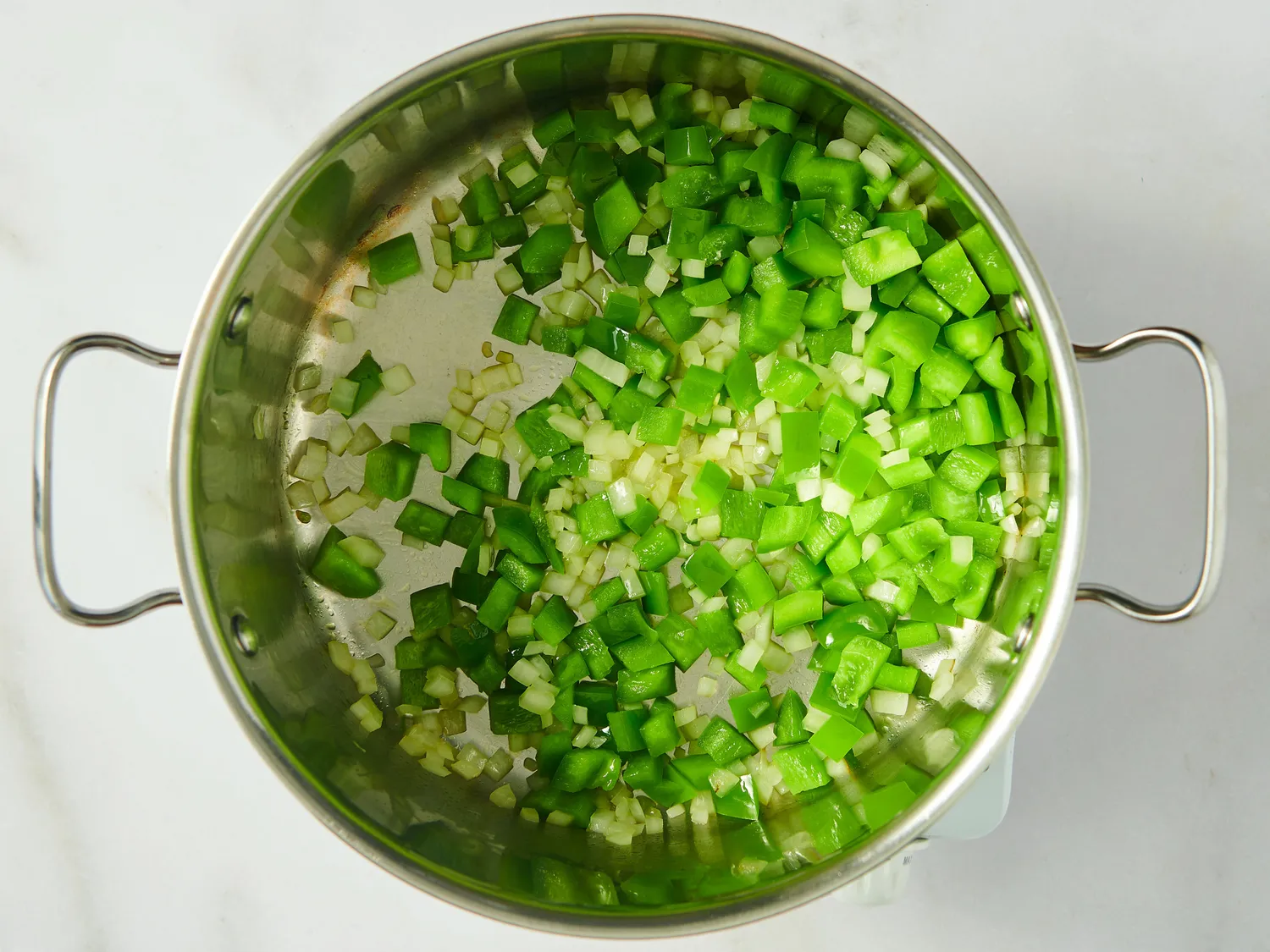 A pot with diced green bell peppers and onions being sautéed on a stovetop