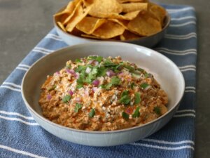 Bowl of mexican street corn dip on blue towel with bowl of tortilla chips in background