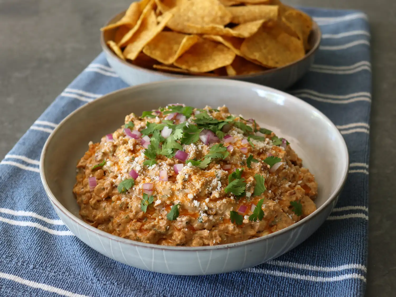 Bowl of mexican street corn dip on blue towel with bowl of tortilla chips in background