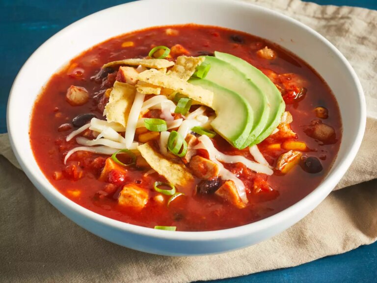 A low angle, close up view of a single bowl of chicken tortilla soup topped with shredded cheese, crispy tortilla strips, and slices of avocado.