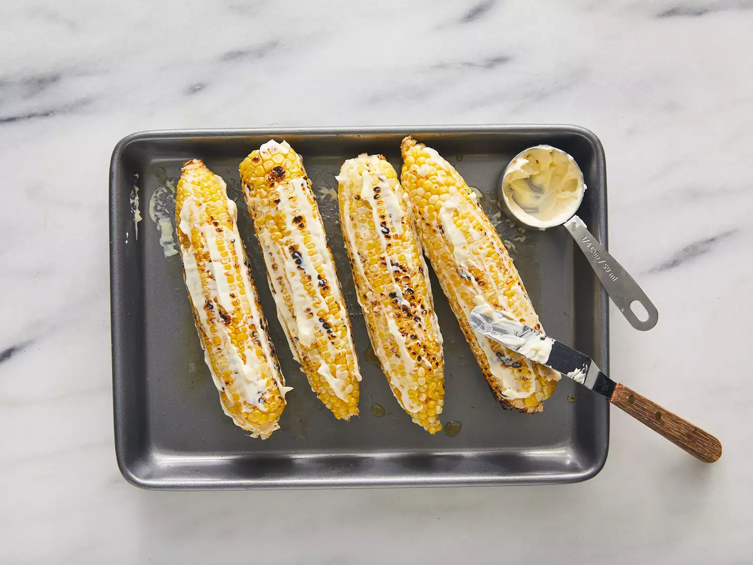 Overhead of cooked Elote being covered in butter on a baking sheet. 
