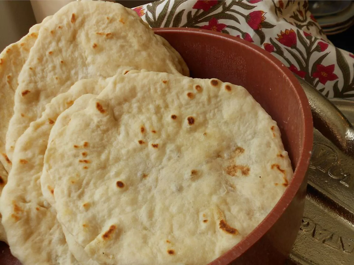 Homemade flour tortillas in a red bowl with a floral tea towel