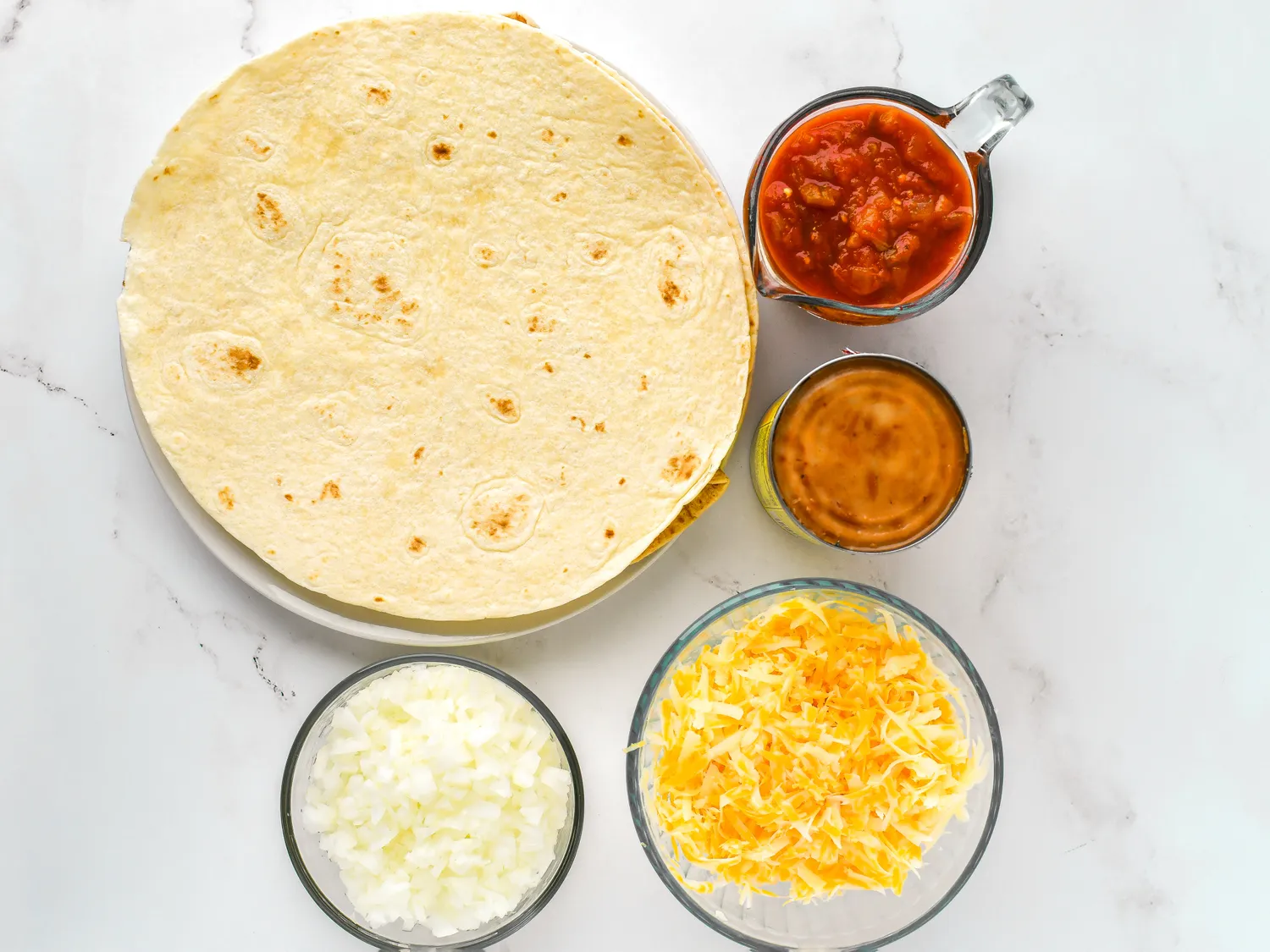 Ingredients for a Mexican casserole arranged on a surface including tortillas shredded cheese diced onions salsa and sauce
