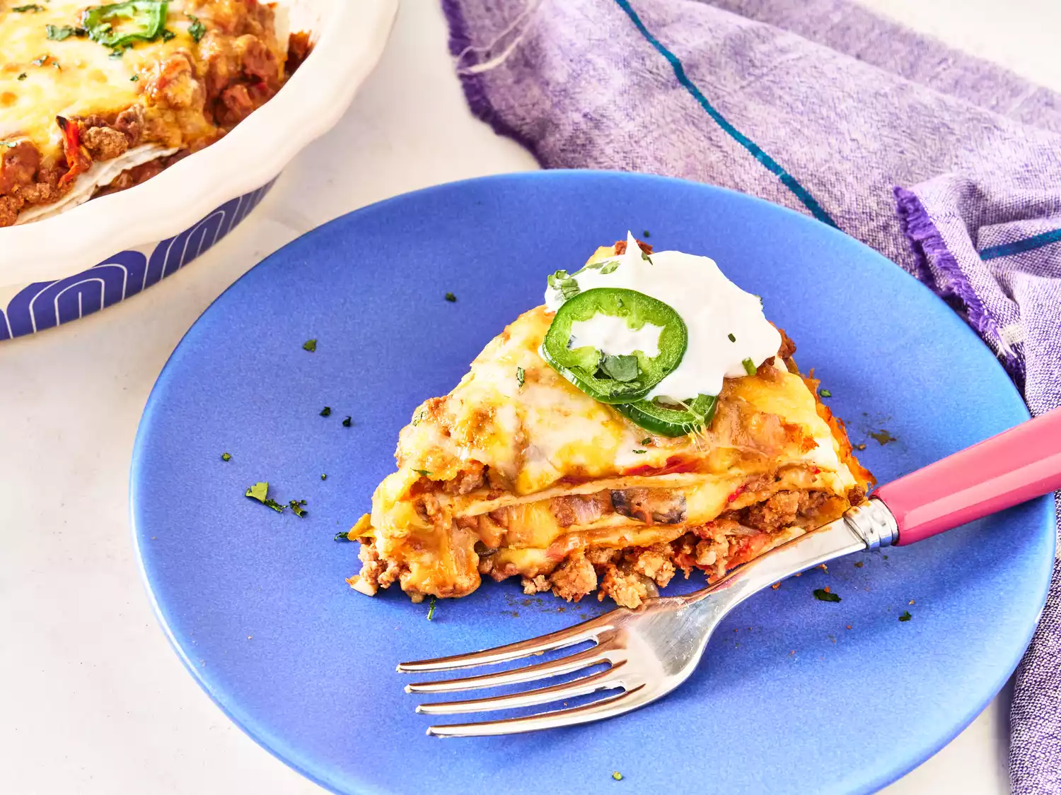 Slice of burrito pie garnished with jalapeño and sour cream on a blue plate with a fork