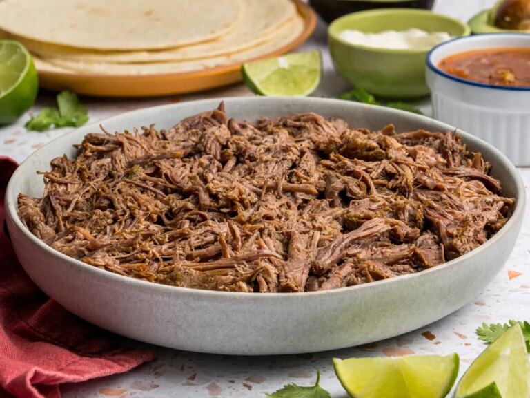 A bowl of slowcooked shredded meat with tortilla wraps and garnishes in the background