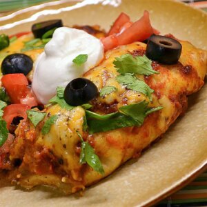 Close up view of beef enchiladas with spicy red sauce garnished with sour cream, black olives, and fresh herbs on a plate