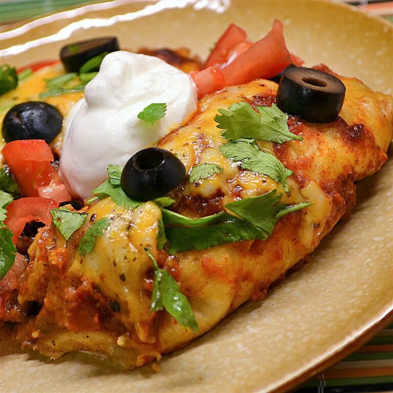 Close up view of beef enchiladas with spicy red sauce garnished with sour cream, black olives, and fresh herbs on a plate
