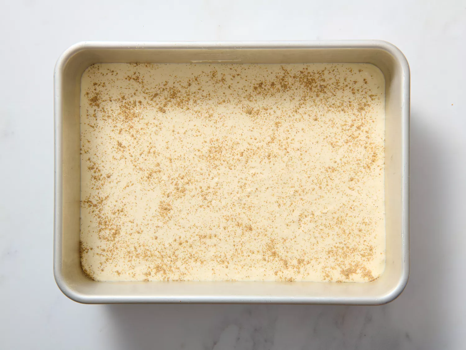 A rectangular pan containing a creamy dessert mixture, sprinkled with spices on top, prepared for refrigerating or baking