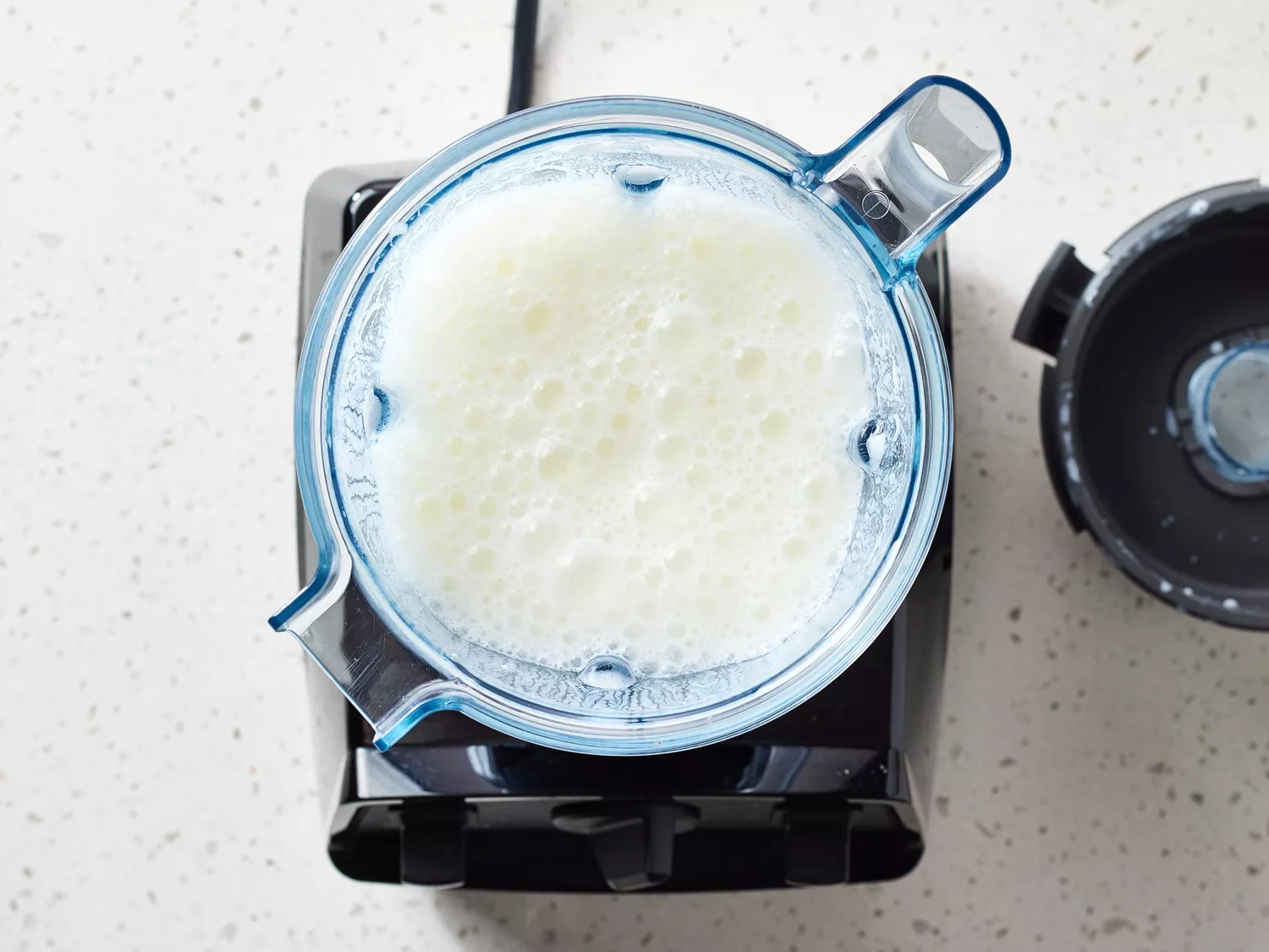 A blender containing a foamy liquid viewed from above, with the lid placed nearby on a countertop