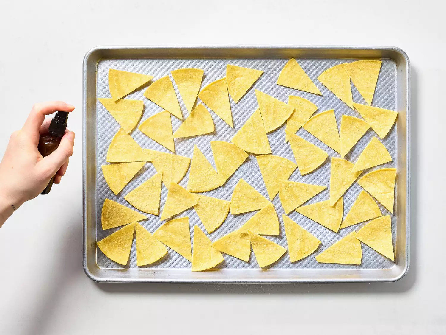Hand spraying oil onto tortilla wedges on a baking sheet.