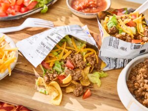 Opened taco in a bag filled with taco ingredients on wooden board next to different taco ingredients