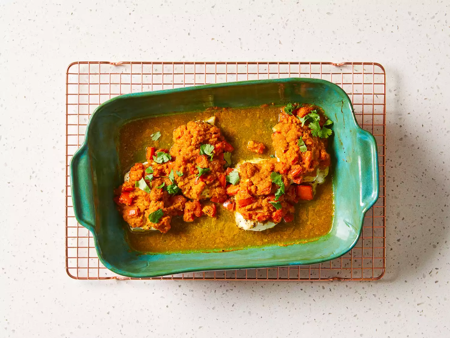 Baked fish fillets topped with tomato and herb sauce in a green baking dish