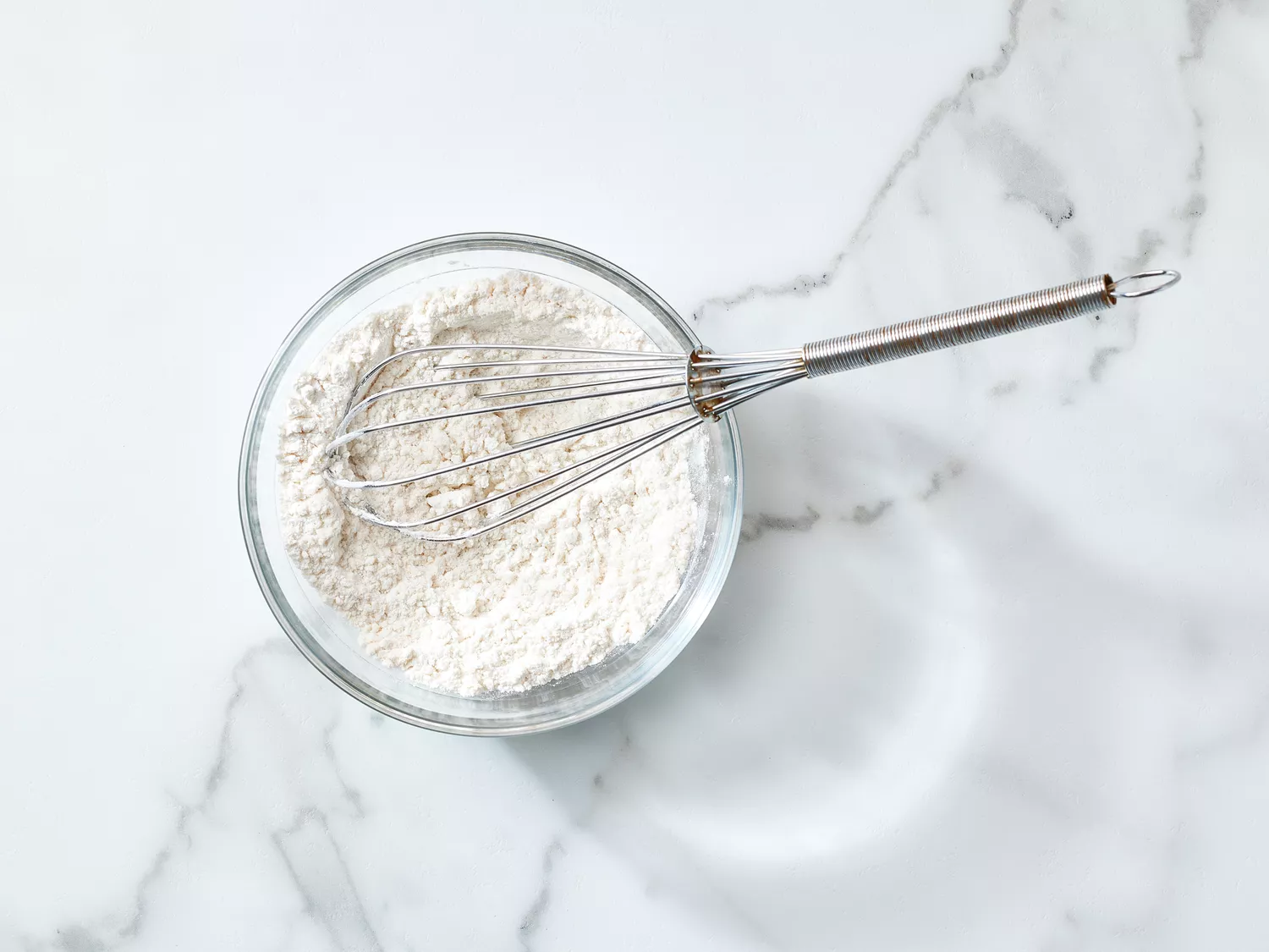 A bowl of flour with a whisk, placed on a marble surface