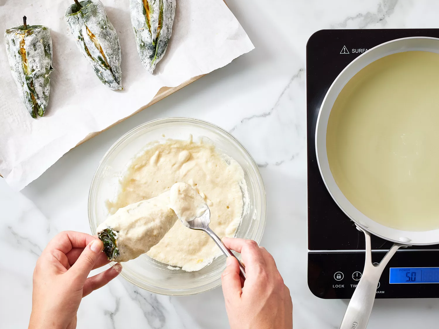 Hands coating chilies with batter near a stovetop and frying pan, preparing chiles rellenos