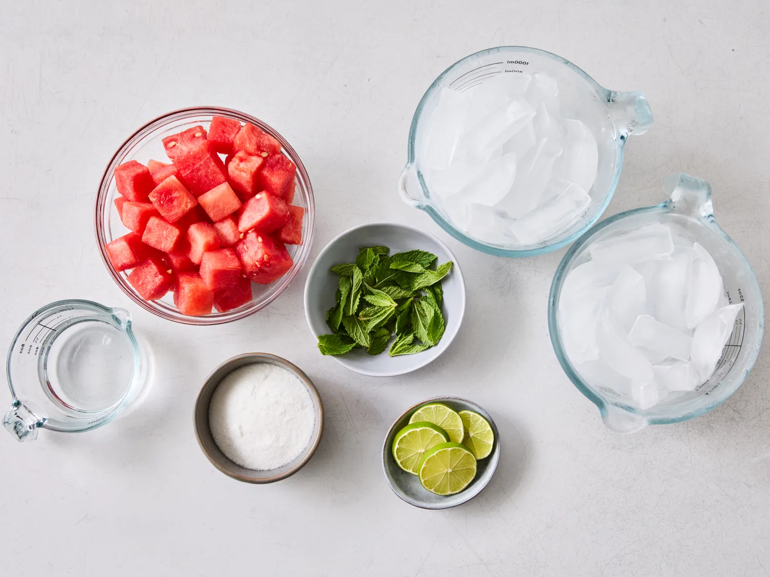 Ingredients for watermelon agua fresca arranged on a surface; includes watermelon, lime slices, mint, ice cubes, sugar, and water