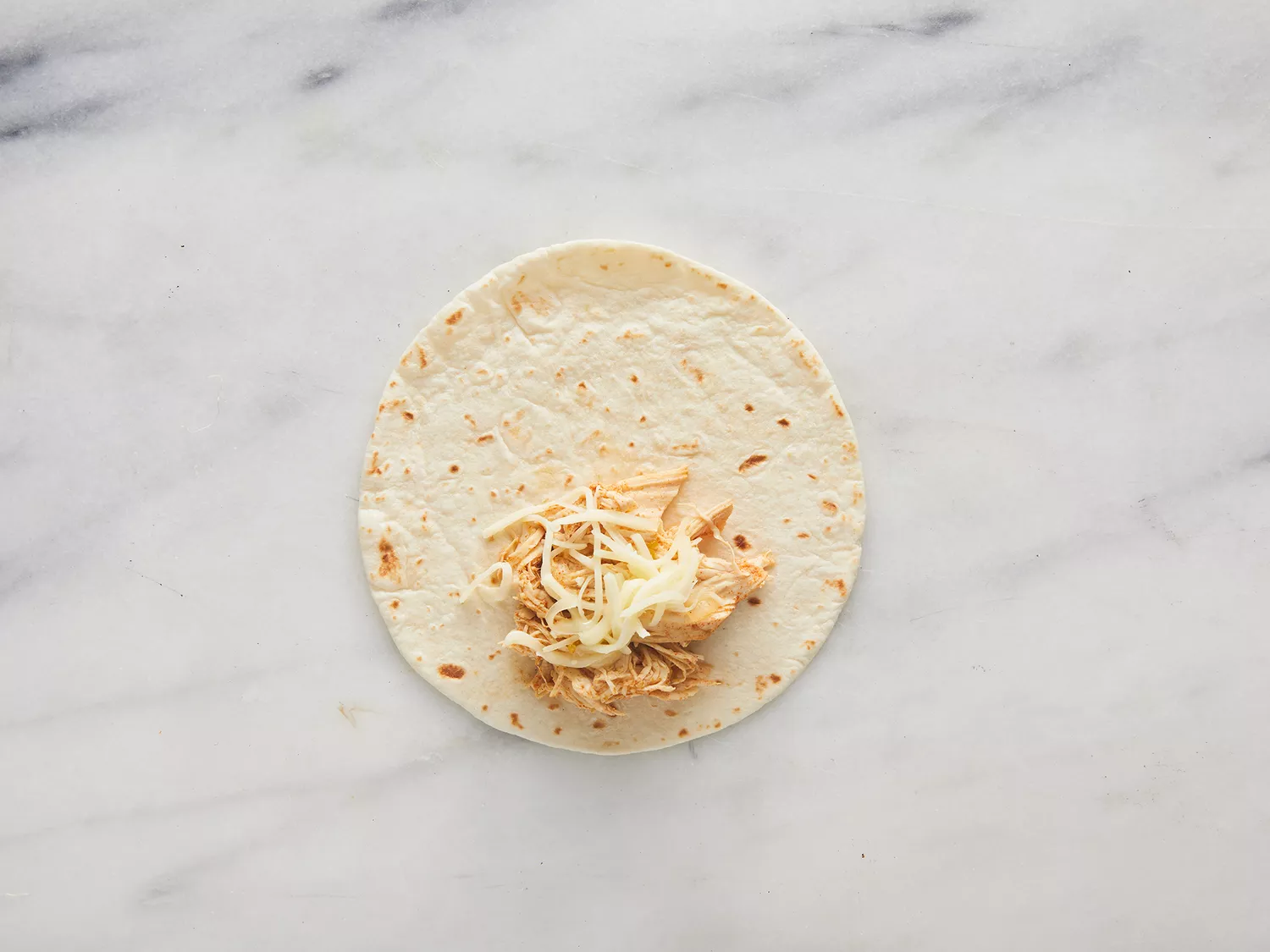 Overhead of shredded chicken and cheese resting on a tortilla. 