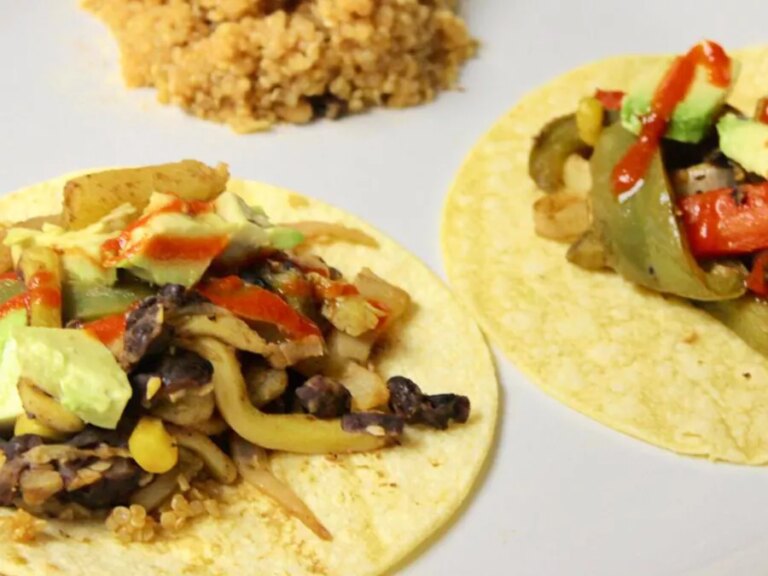 Close up view of vegan fajitas with peppers, avocado and red sauce on corn tortillas, served with grains on a white plate