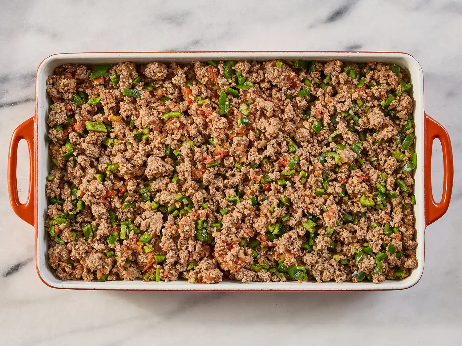 A baking dish filled with a mixture of ground meat and chopped vegetables prepared as a step in making a hot tamale pie