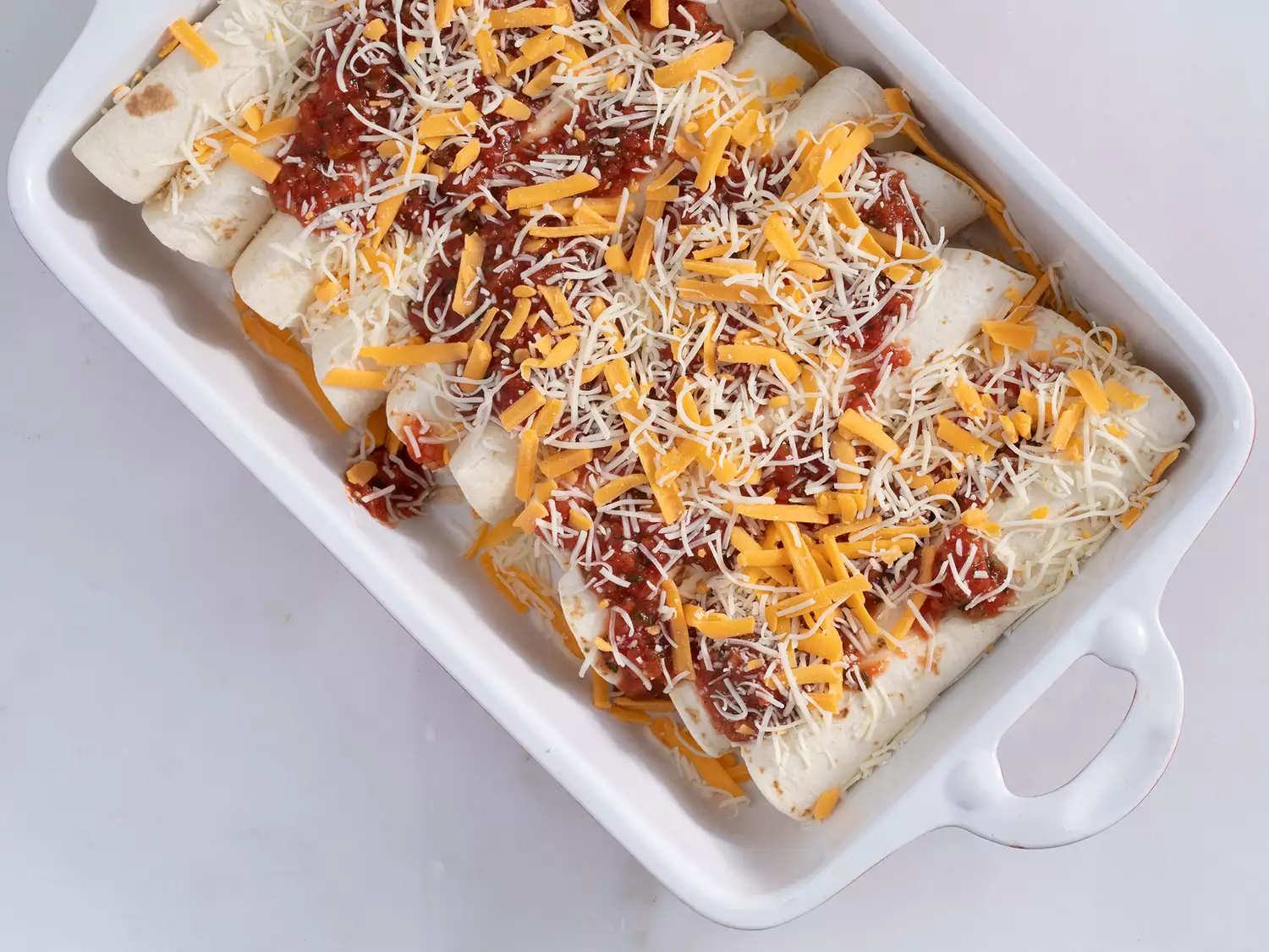 Rolled enchiladas in baking dish with cheese mixture, salsa, and green onions spread on top