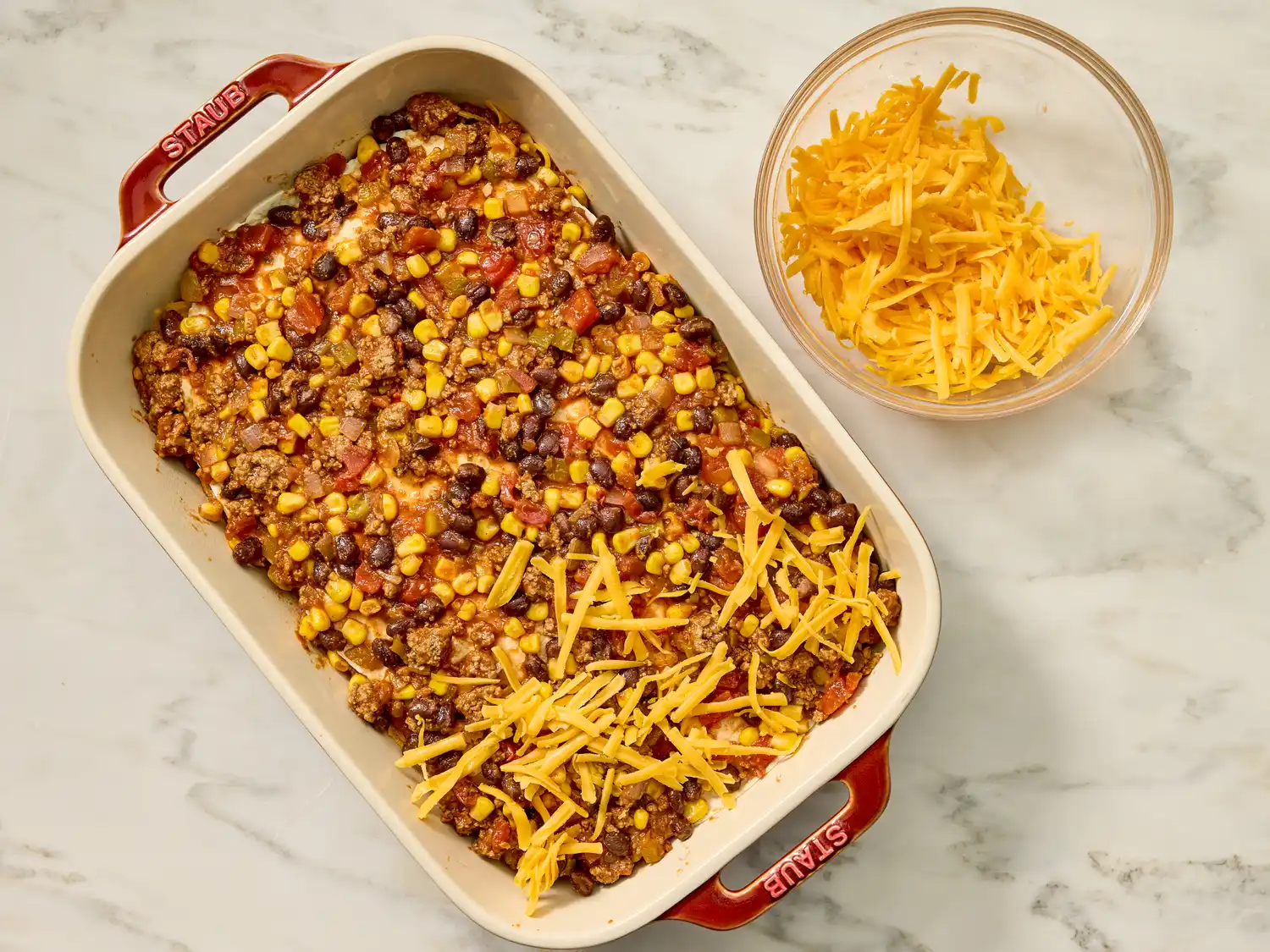 Casserole dish with layered ingredients including ground meat beans corn and shredded cheese a bowl of cheese beside