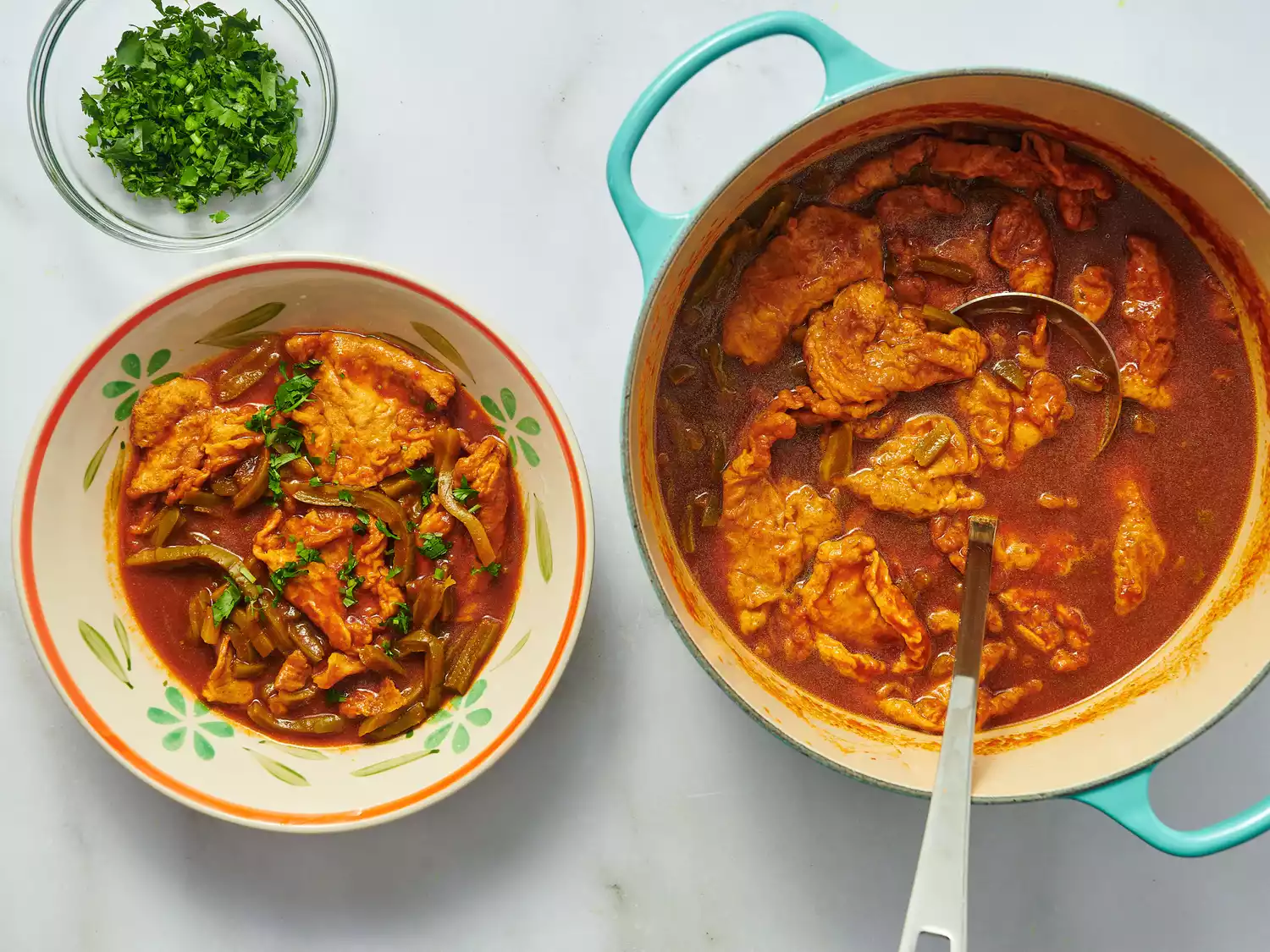 A pot and bowl both containing tortas de camaron in red sauce, garnished with herbs. A small bowl of chopped herbs is beside them