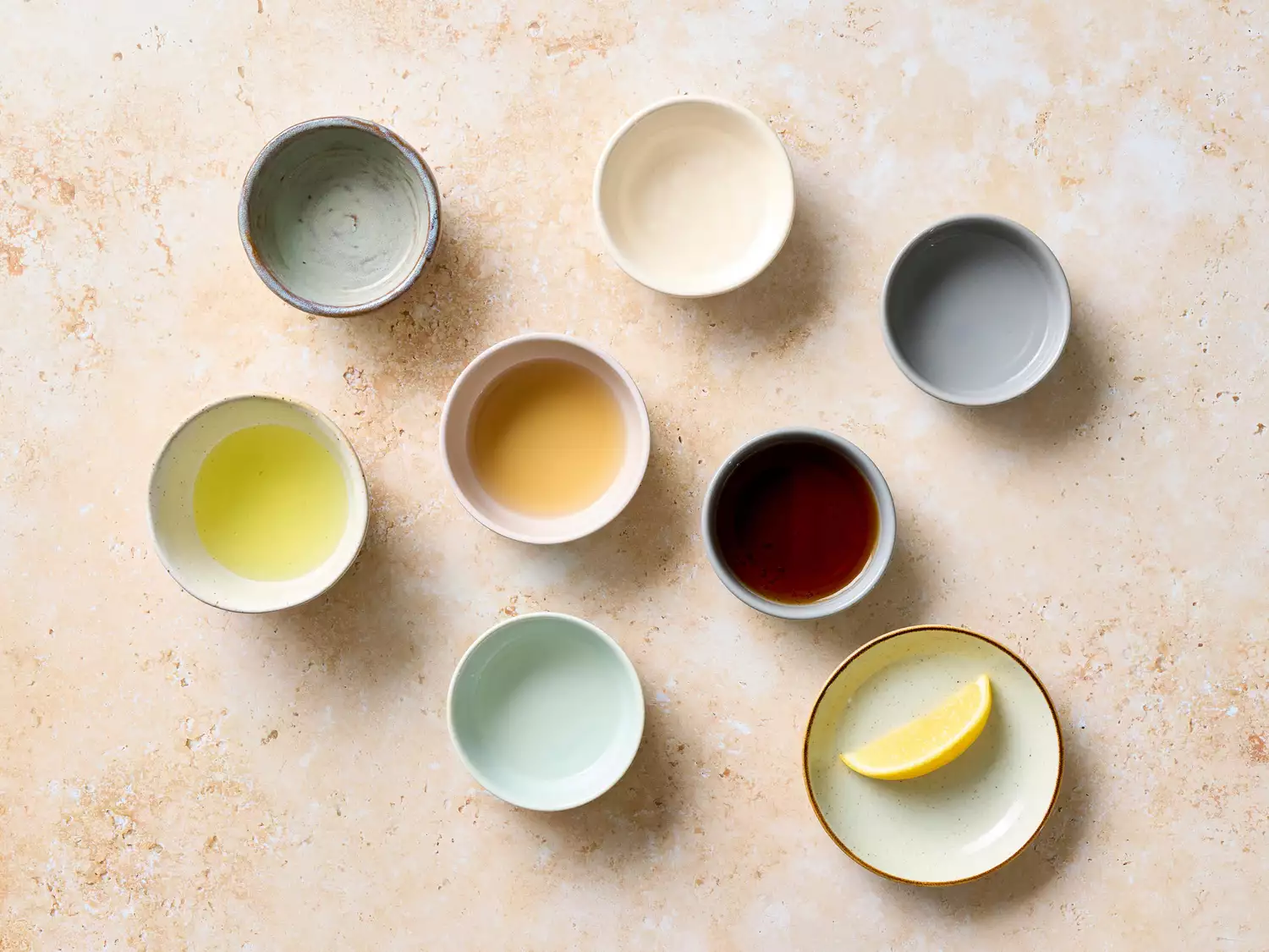Assorted ingredients for a cocktail on a beige surface, including lemon wedge, syrups, and liquids in small bowls.