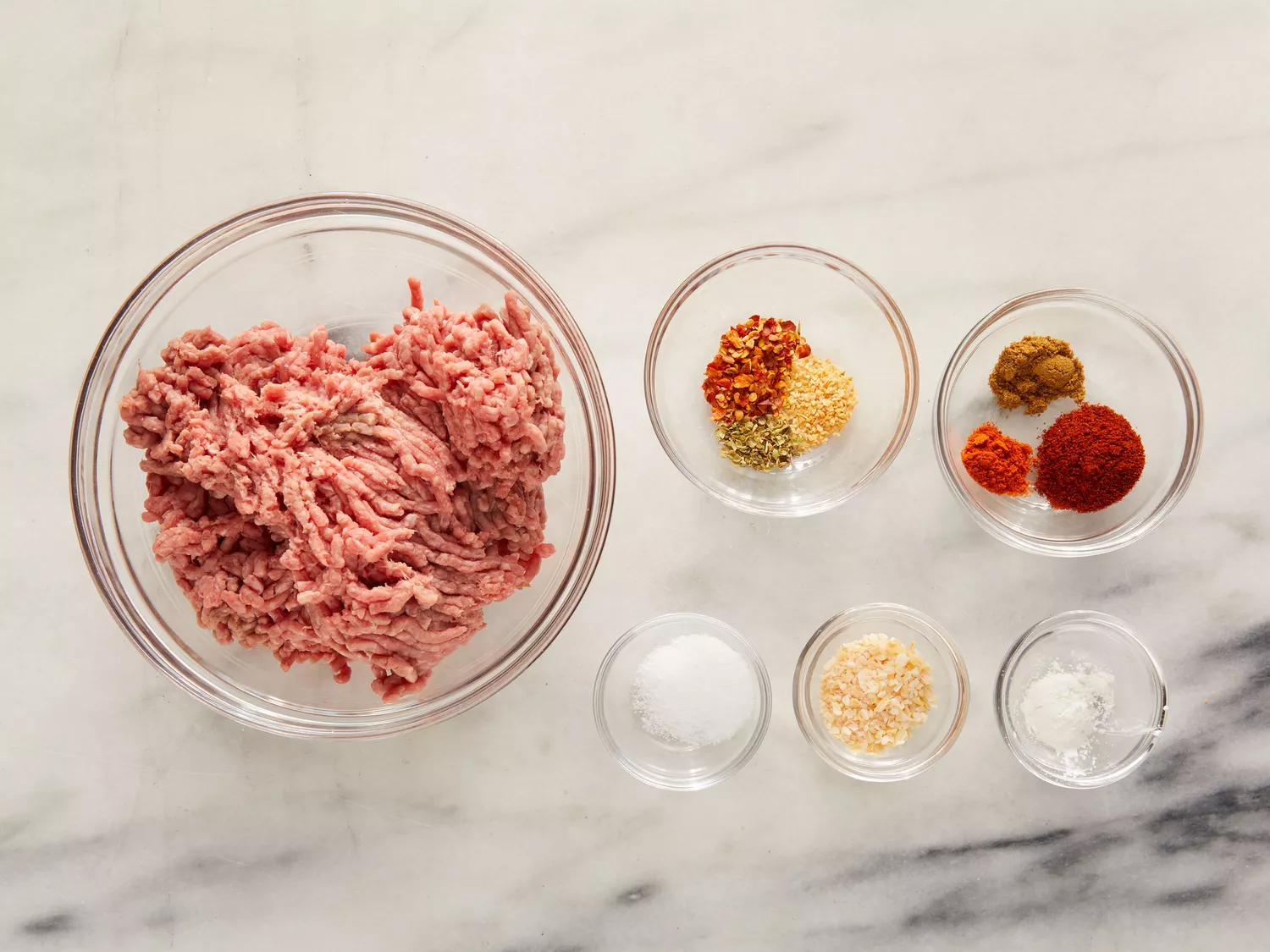 Bowl of raw ground beef and smaller bowls with seasoning mixes for taco seasoning placed on marble counter