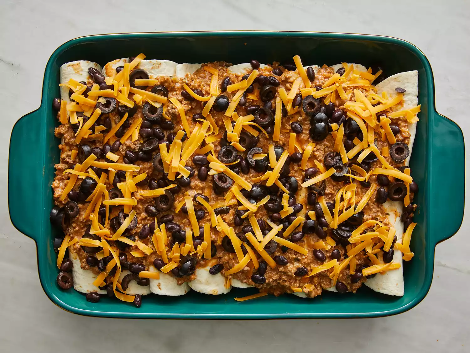 a top down view of an unbaked enchilada casserole topped with sauce, sliced black olives, and shredded cheddar cheese. 