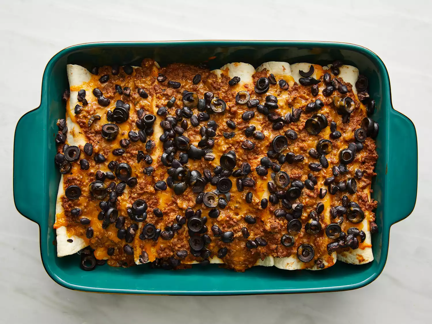  a top down view of baked beef enchiladas in a turquoise 9x13 casserole . 