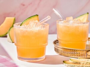 Two glasses of aqua fresca with ice and melon slices, a gold tray, on a marble surface