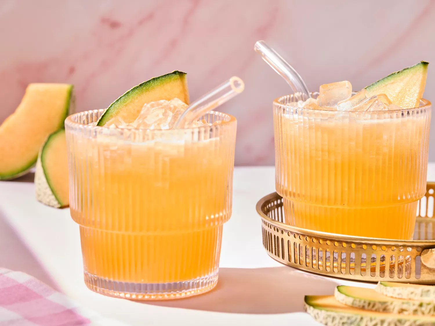 Two glasses of aqua fresca with ice and melon slices, a gold tray, on a marble surface