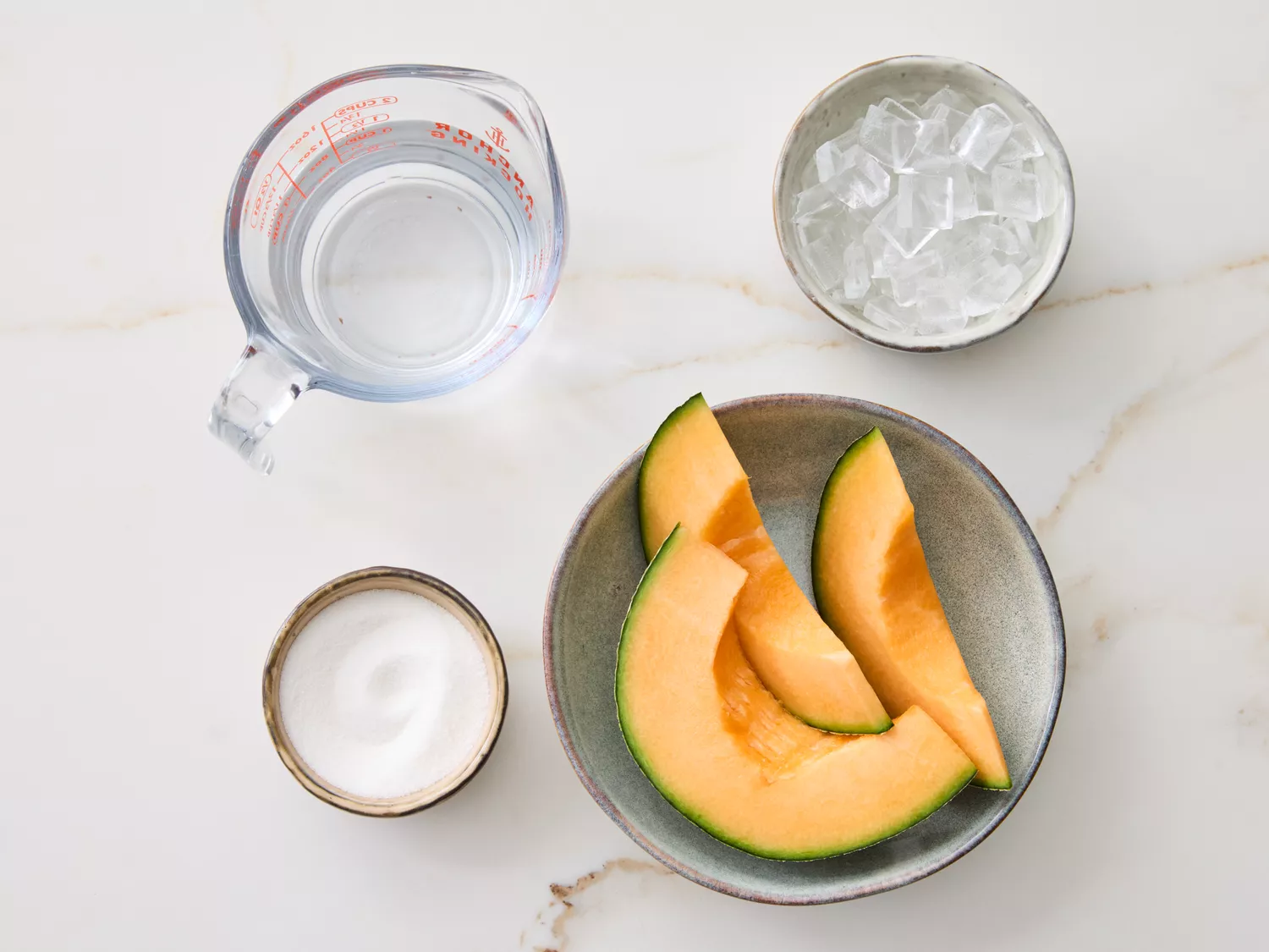 Four ingredients: melon slices, a bowl of ice, a measuring cup with water, and a small bowl with a white ingredient on a marble surface
