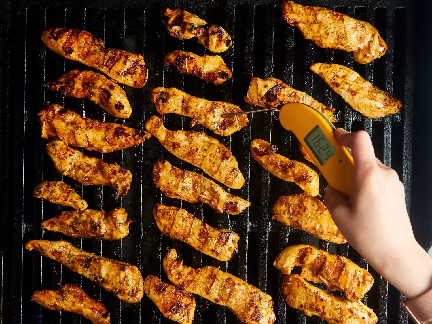 Grilled chicken strips being cooked while a hand checks their temperature with a thermometer