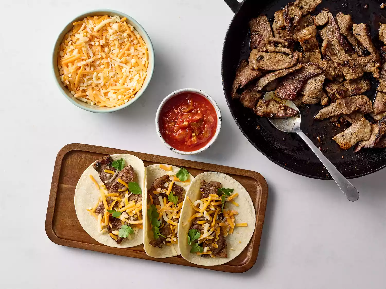 Assembled beef fajitas with cheese and salsa on a tray next to beef in a skillet and bowls with cheese and salsa 