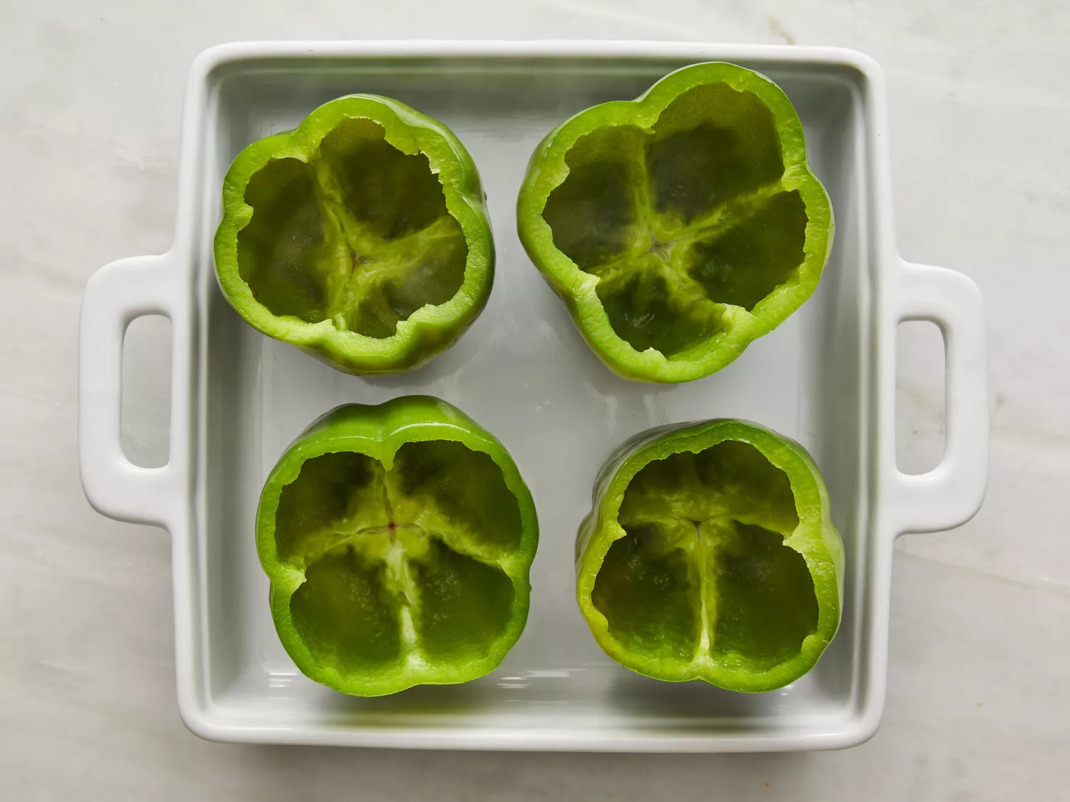 Overhead of four green bell peppers with their tops cut off and resting in a baking dish. 