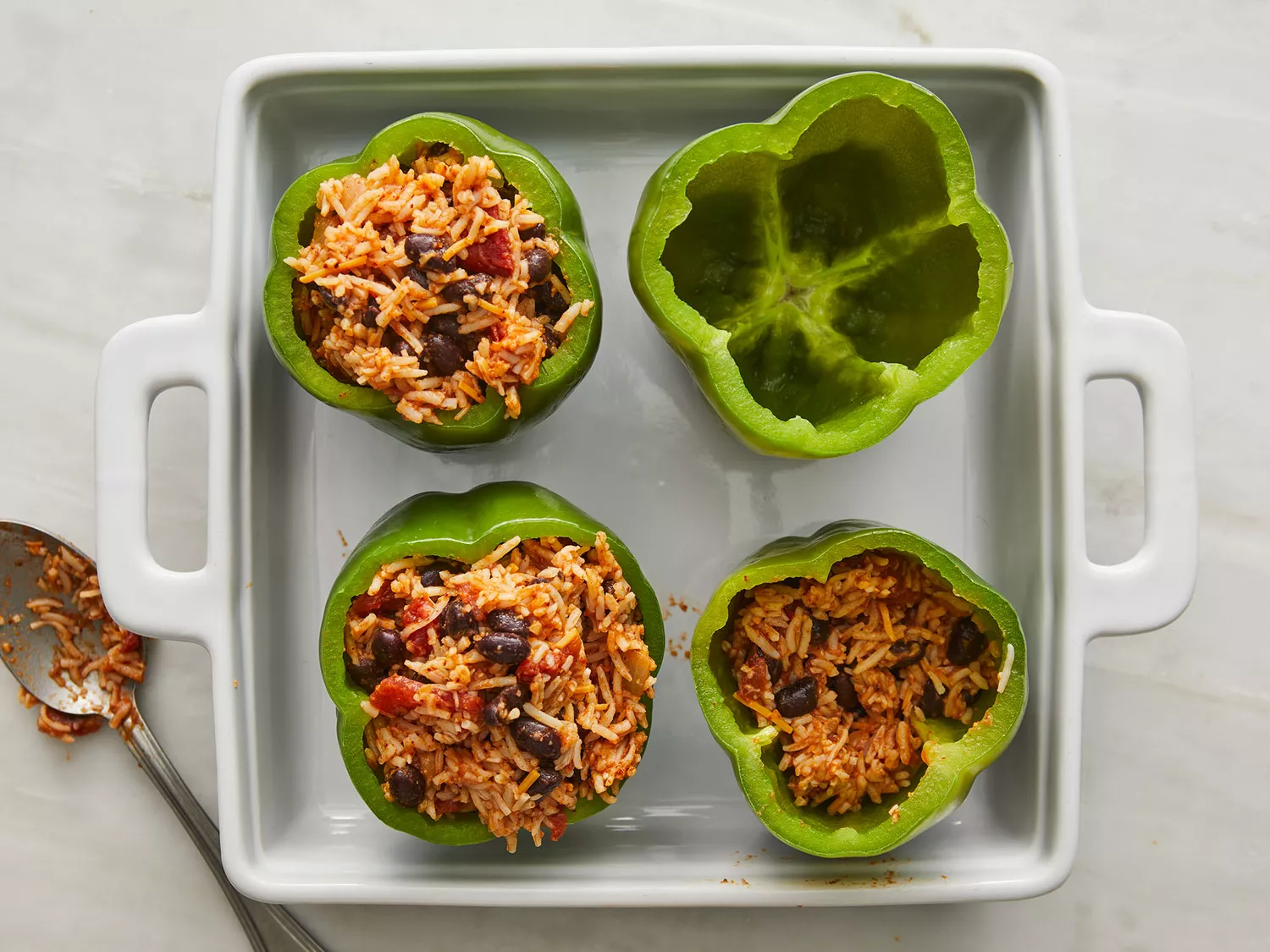 Overhead of a cheese and rice mixture being spooned into four green bell peppers as they rest in a baking dish. 