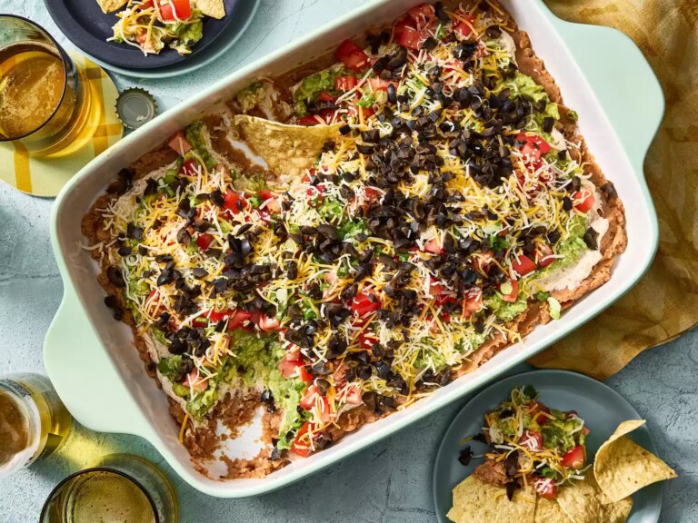 Baking dish with seven layer dip topped with cheese, olives, and tomatoes.