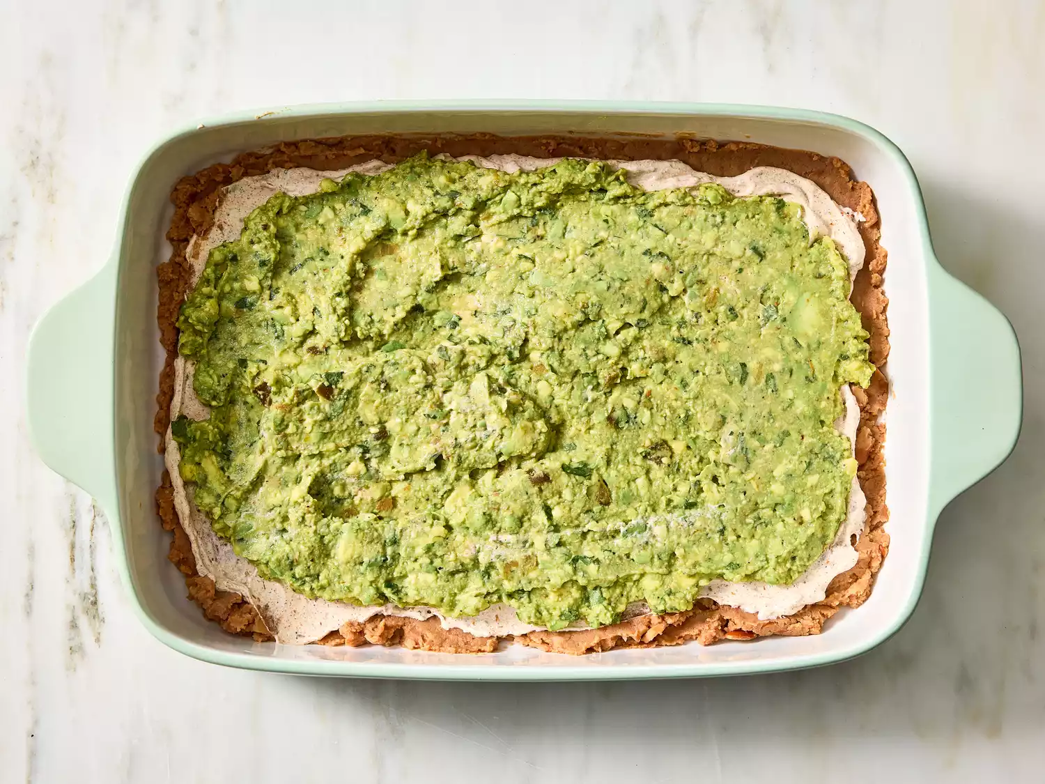Layer of guacamole spread over bean dip in baking dish.