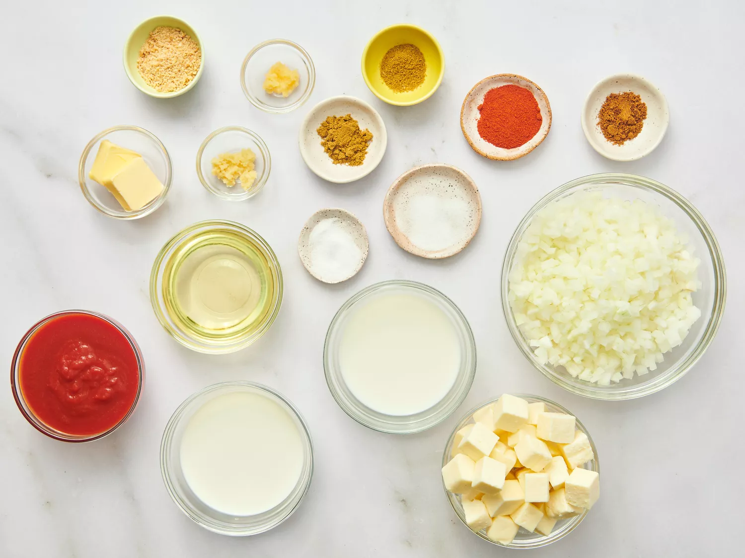 Prepared ingredients for paneer butter masala laid out in bowls on a white surface, including cream, spices, chopped onions, and cubes of paneer