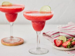 Two frozen strawberry margaritas garnished with lime slices, with strawberries and lime on a plate nearby