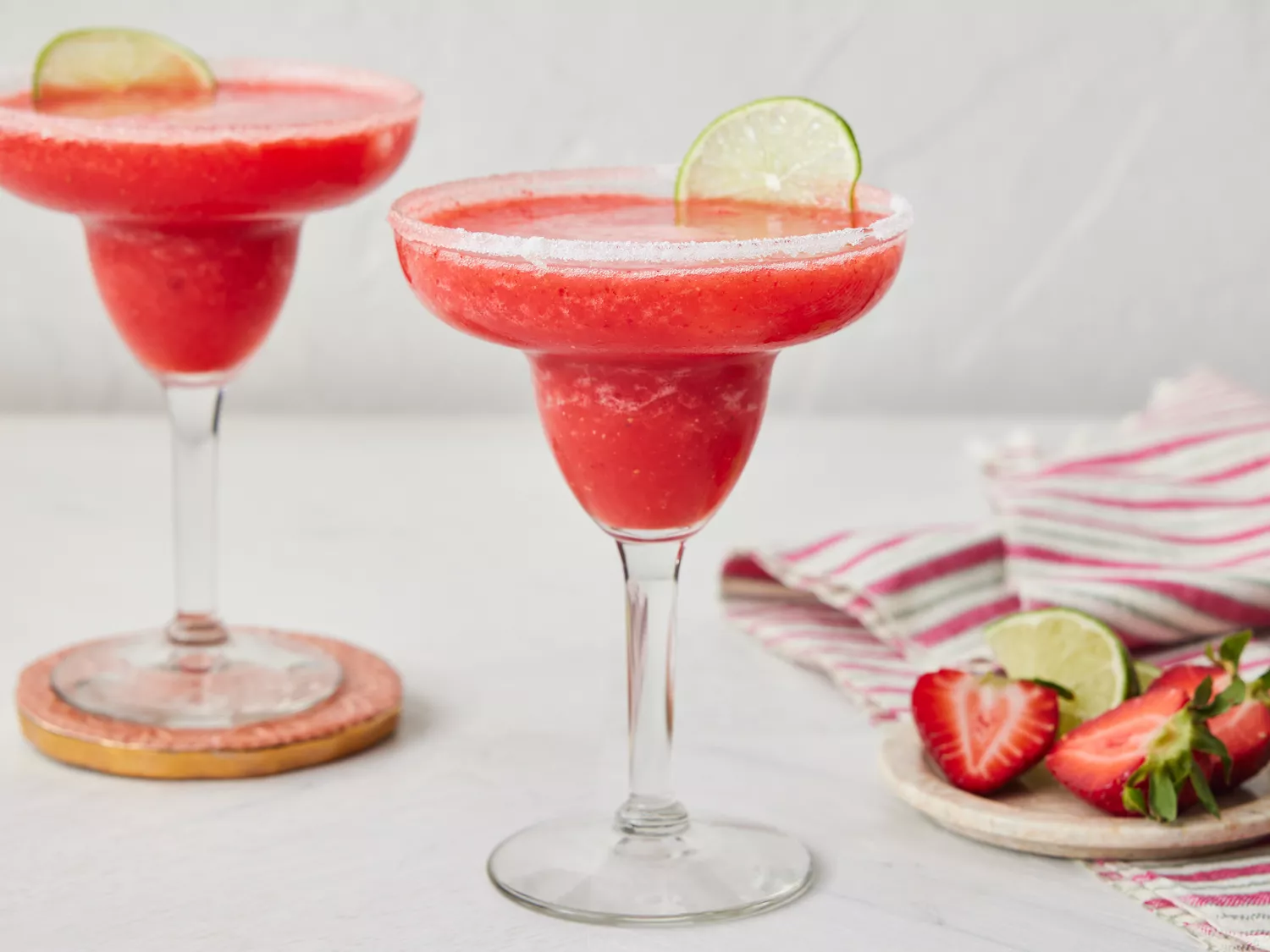 Two frozen strawberry margaritas garnished with lime slices, with strawberries and lime on a plate nearby